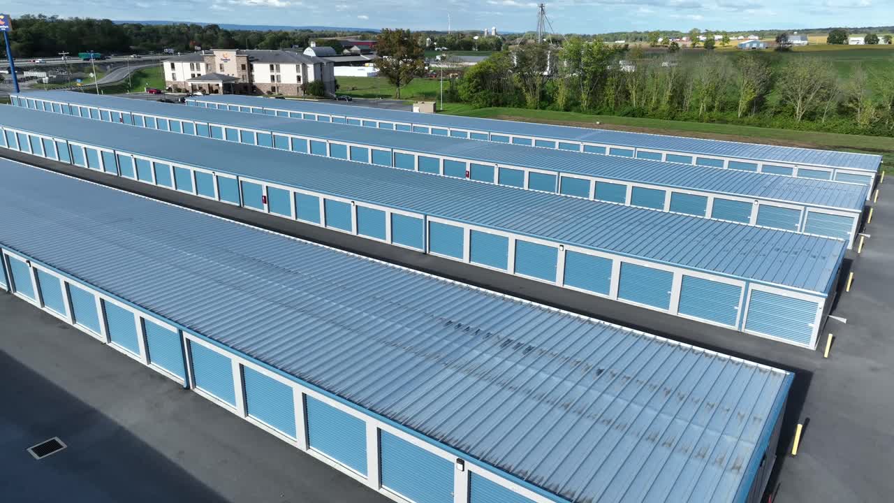 Blue rental warehouse garages and self storage parking in American suburbia. Aerial orbit wide shot. Traffic on highway in fall season