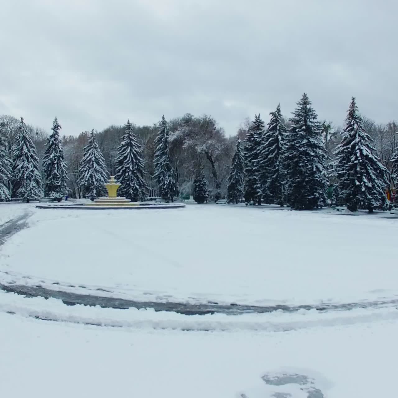 Square with non-working fountain covered with white snow. Beautiful fir-trees surrounding the square in the urban park. Drone footage