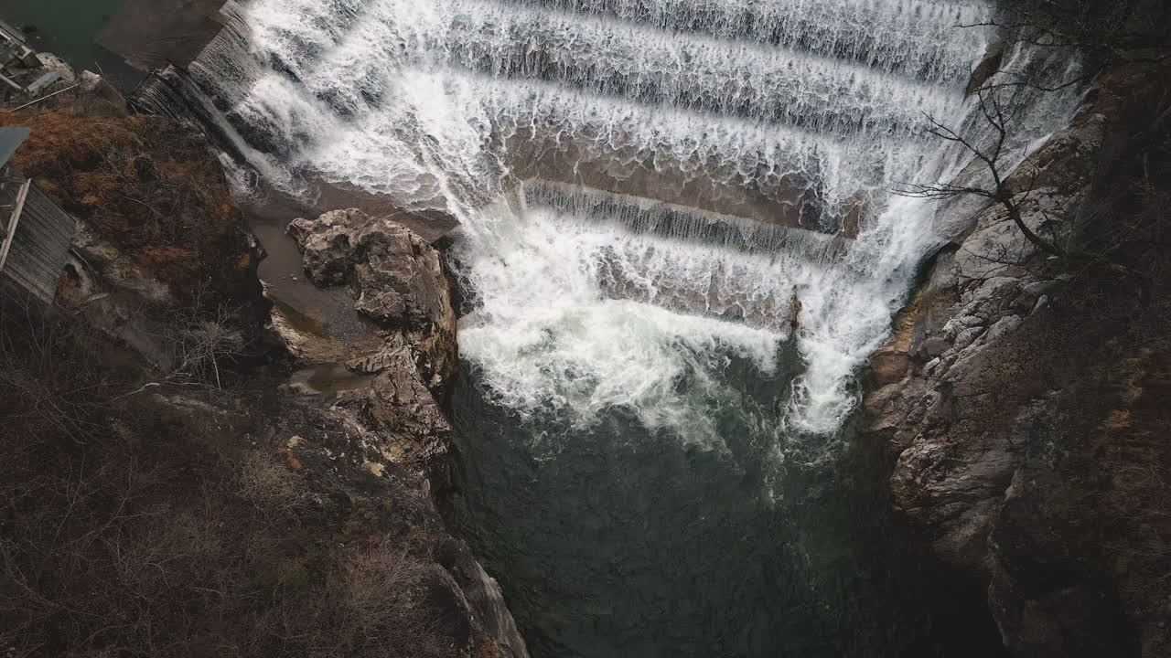 Drone glides down to Lechfall dam's tiered spillway in Fussen, capturing powerful water flow into gorge pool surrounded by autumn foliage and rocks. Dynamic hydro reveal