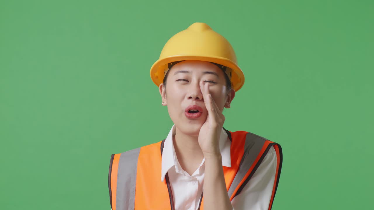 안전 헬을 입은 아시아 여성 엔지니어가 녹색 스크린 배경 스튜디오에 서 있는 동안 입 위에 손으로 소리치고 있습니다.