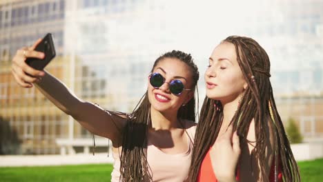 happy women with dreads sitting on grass in summer park and talking selfies. young friends talking and taking photos, posing