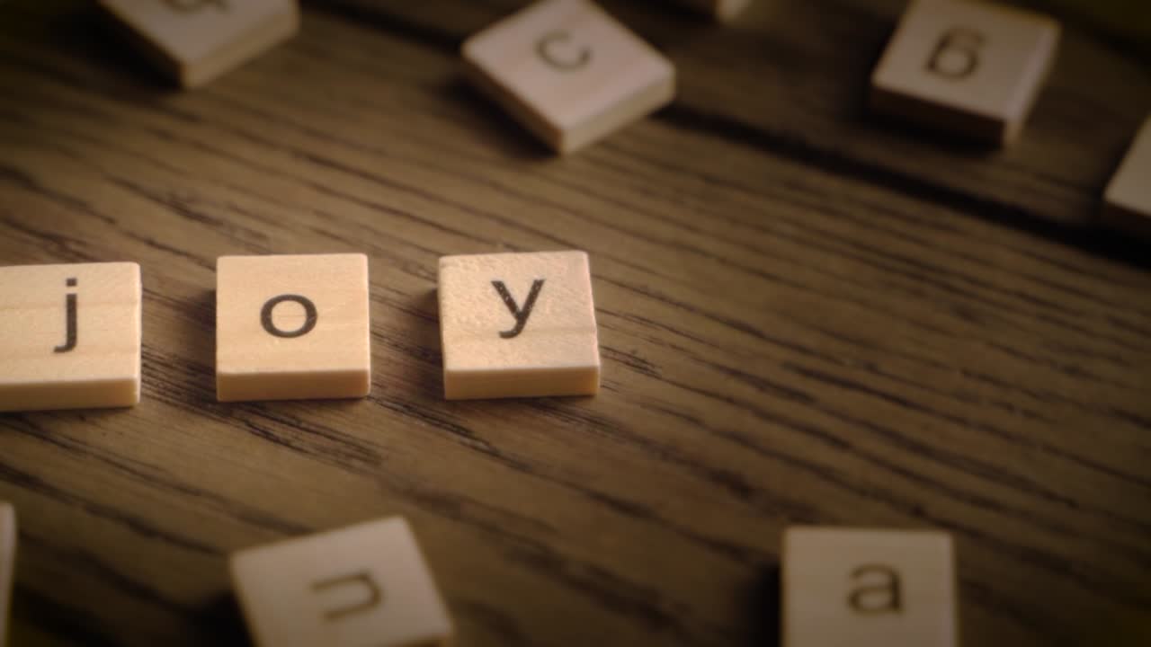 Wooden scrabble-style tiles spell joy as the camera pans smoothly across a rustic wooden surface with warm lighting.