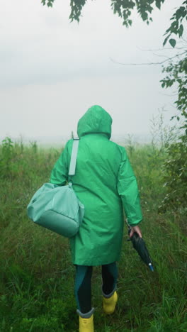 vista trasera de una mujer con un impermeable verde que lleva una bolsa de color menta y un paraguas mientras usa botas de lluvia amarillas, caminando por un camino rodeado de vegetación y árboles