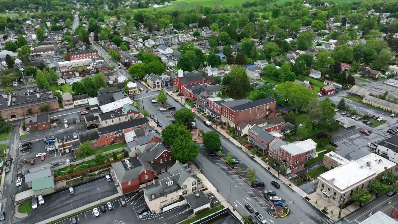 Establishing drone shot of american city with historic Buildings and flag of USA. Green trees during cloudy spring day. Descend aerial wide shot. Parking cars in center of city. Manheim,PA, USA.