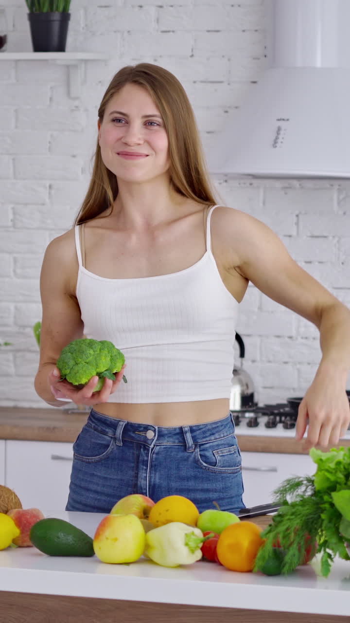 Attractive woman at the table with fresh vitamins. Beautiful sportswoman in casual wearing holding broccoli and shows her arm muscles in the kitchen. Healthy lifestyle. Vertical video