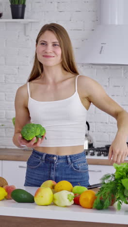 attractive woman at the table with fresh vitamins. beautiful sportswoman in casual wearing holding broccoli and shows her arm muscles in the kitchen. healthy lifestyle. vertical video