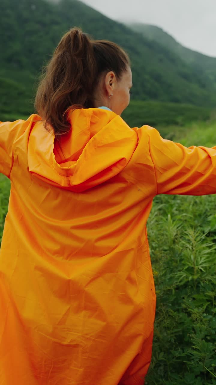 Woman in Orange Raincoat Walking Through Lush Green Mountain Field