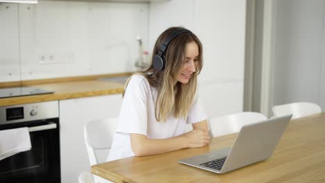 Blonde-woman-make-video-call-online-using-laptop-and-headphones-at-kitchen