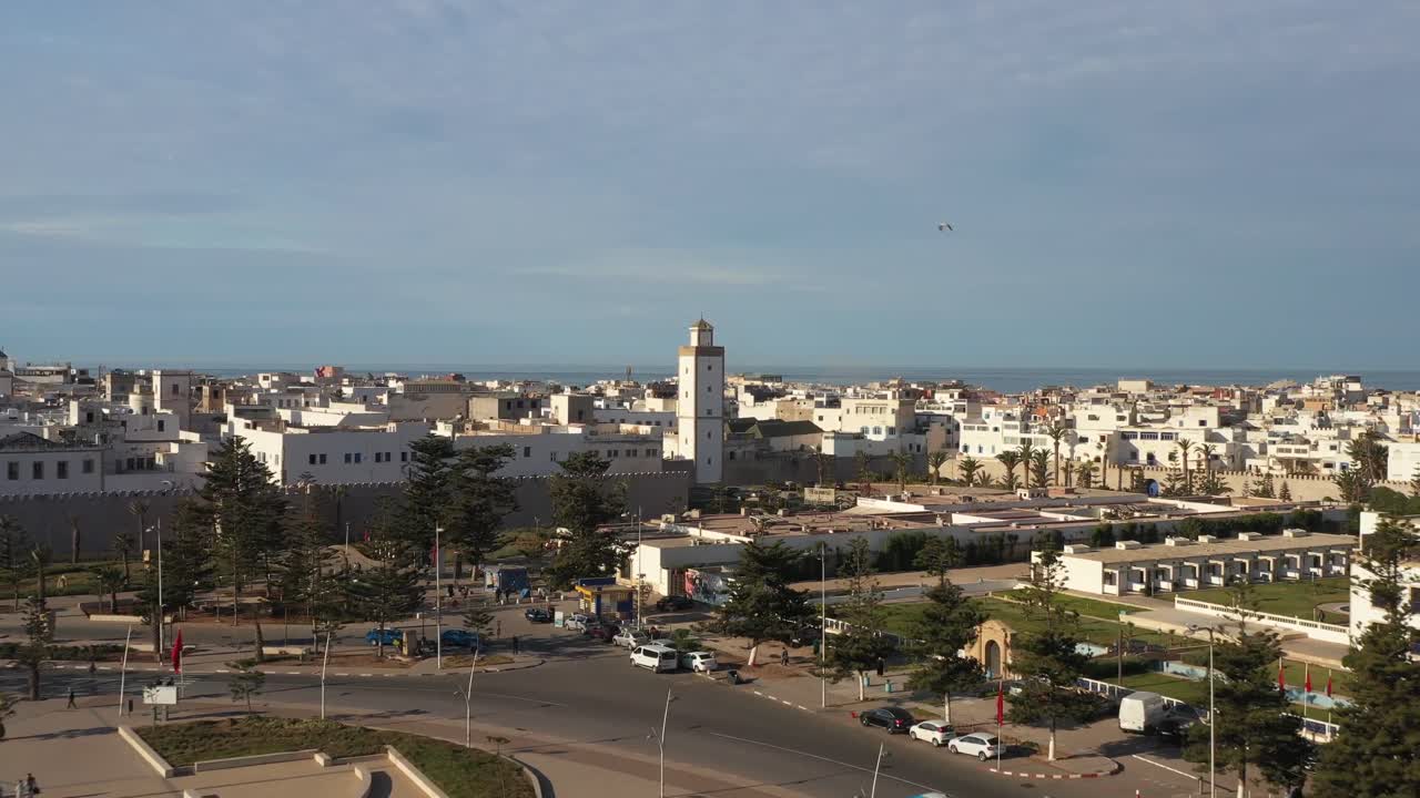 Bredt dronebillede med udsigt over sandstranden, gadetrafikken og den traditionelle hvide arkitektur i Essaouiras medina med en fremtrædende moskéminaret