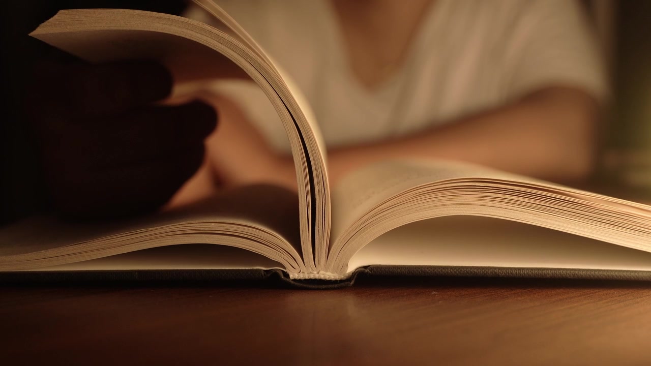 La mano di una donna gira le pagine di un libro aperto appoggiato su un tavolo di legno scuro, immerso nella calda luce dorata di una lampada, creando un'atmosfera di lettura intima e accogliente con un morbido bokeh sullo sfondo.