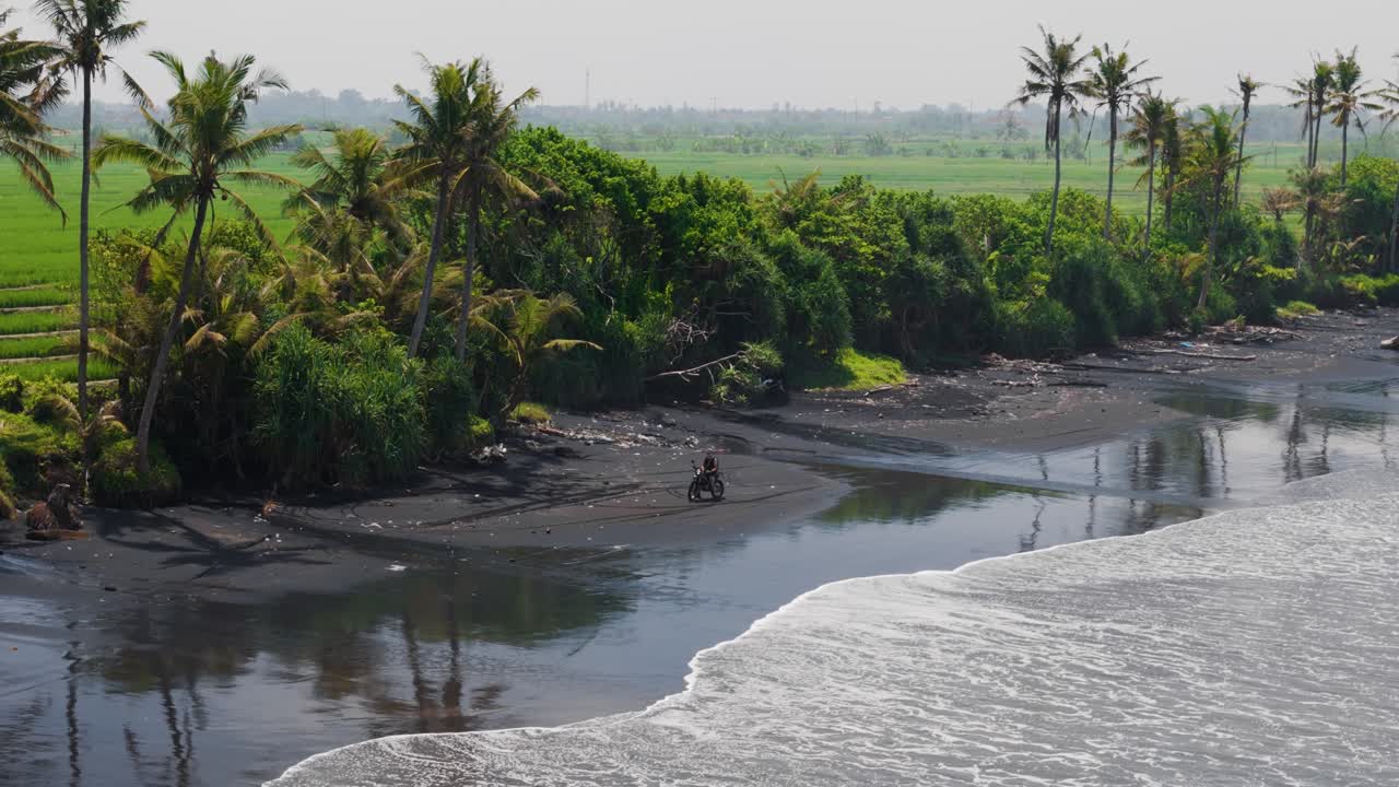 Wide angle aerial approach toward a dirt bike rider on Bali’s black sand shoreline. Lush palm jungle and vivid rice fields frame the rugged coastline in warm tropical light