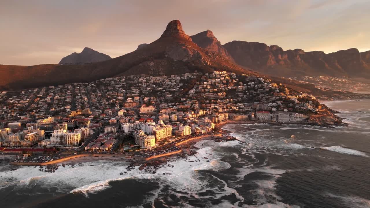 This captivating aerial footage showcases a densely populated coastal city nestled between rocky shores and towering mountains. Bathed in the warm glow of sunrise or sunset, the scene highlights