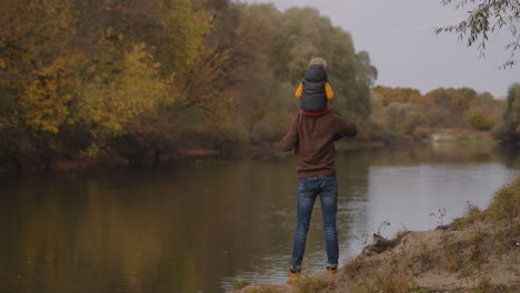 father and little son on shore of river at autumn day toddler is sitting on shoulders of dad rear view family weekend at nature happy parenthood and childhood