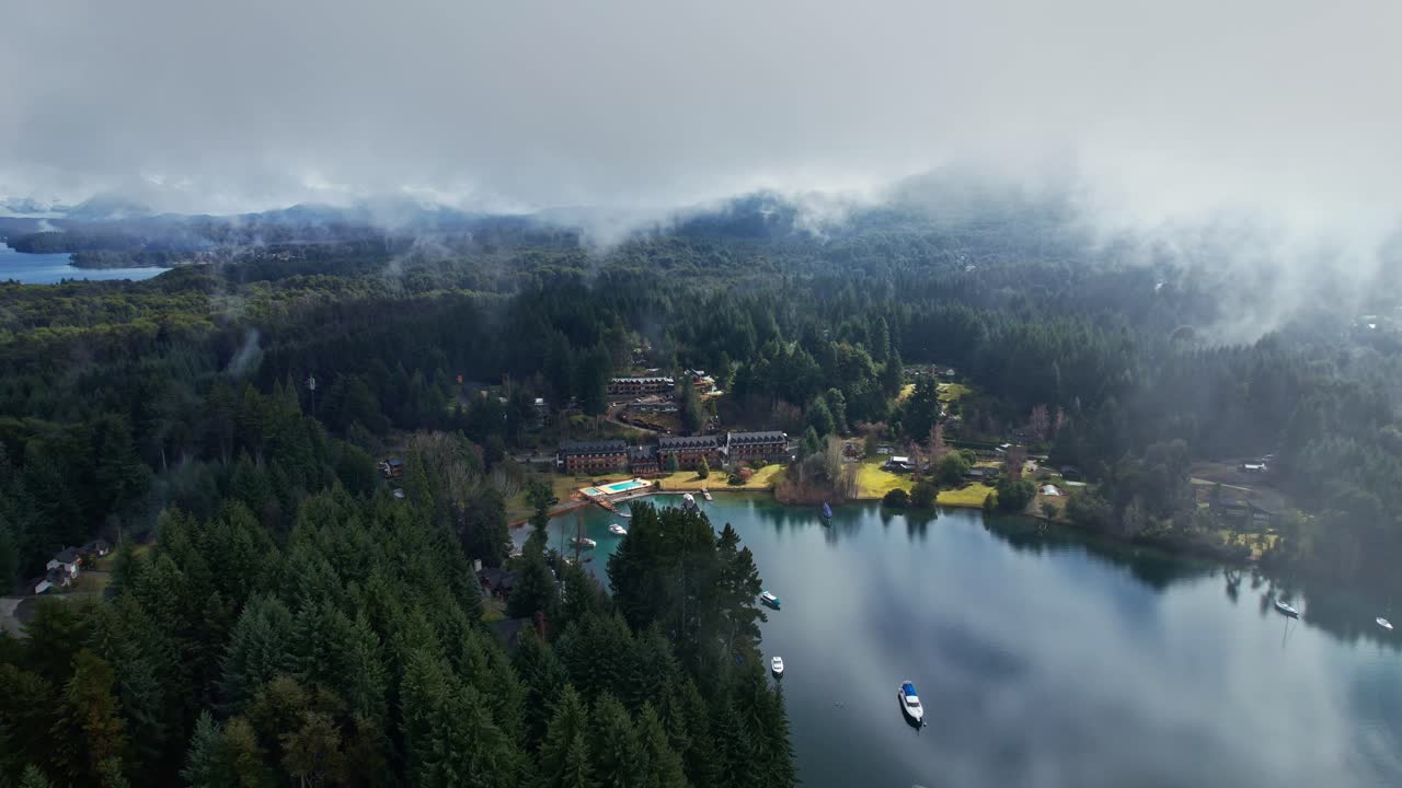 Ripresa in avanti con drone in avvicinamento su un resort turistico a Puerto Manzano, Villa La Angostura. Nuvole basse e nebbia che coprono la pineta e il Lago Nahuel Huapi in Patagonia, Argentina.