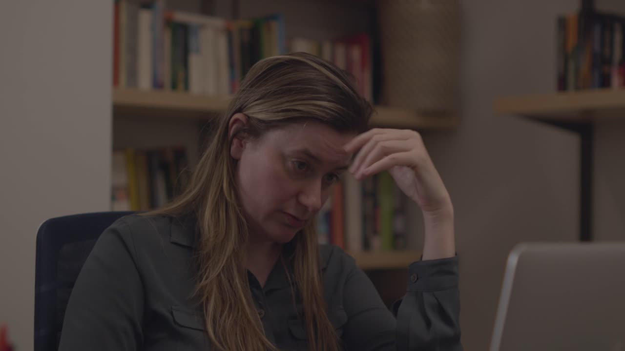 A woman reacts to her laptop with stress, disbelief, and shock. She blows hair out of her face. She sits in an office setting with bookshelves in the background