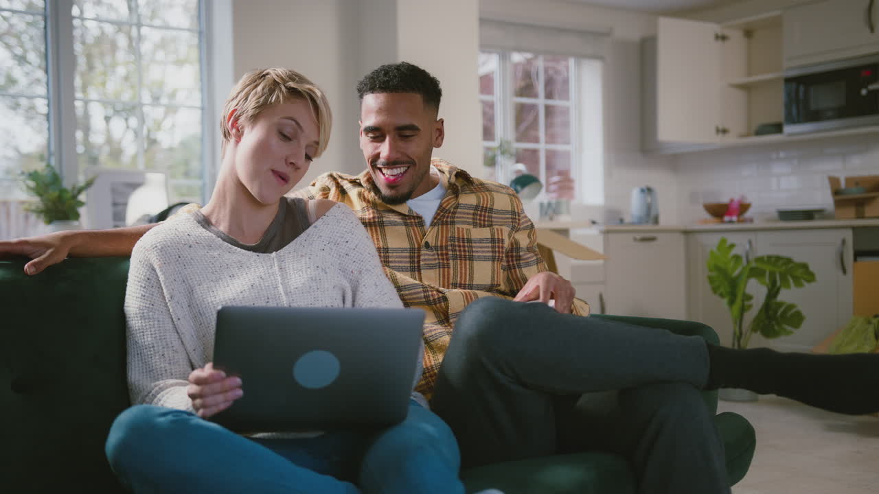 Couple Unpacking Boxes After Moving Into New Home With Laptop And Hot Drinks
