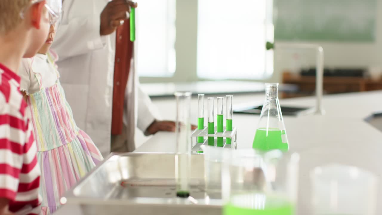 Diverse teacher raising green test tube, guiding students wearing goggles toward rack