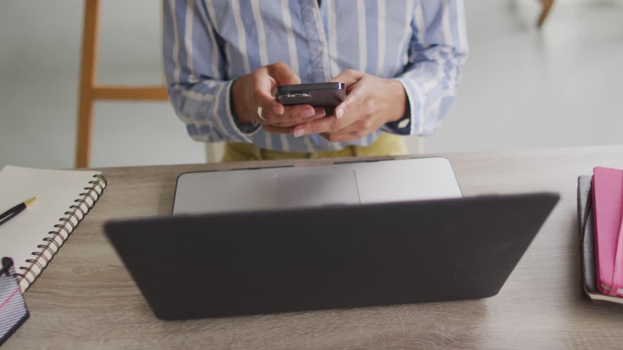 video de la sección media de una diseñadora biracial que trabaja en una computadora portátil y un teléfono inteligente en el estudio
