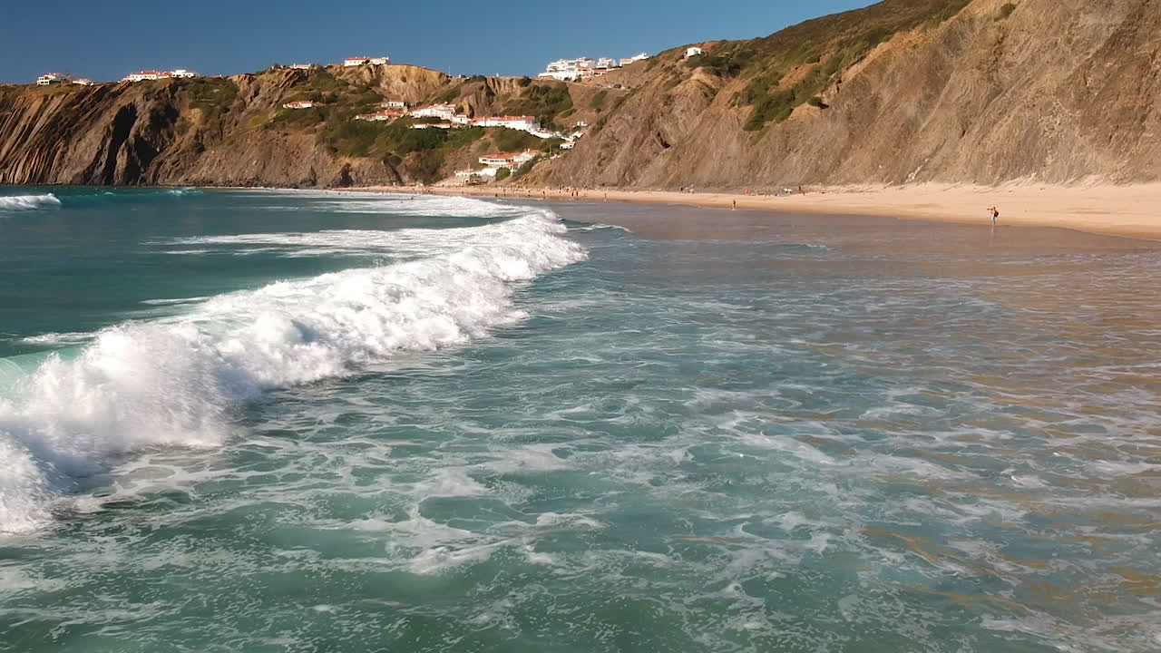 toma aérea de personas nadando en olas de aguas bravas rodando en una playa al lado de casas en un acantilado en algarve, portugal