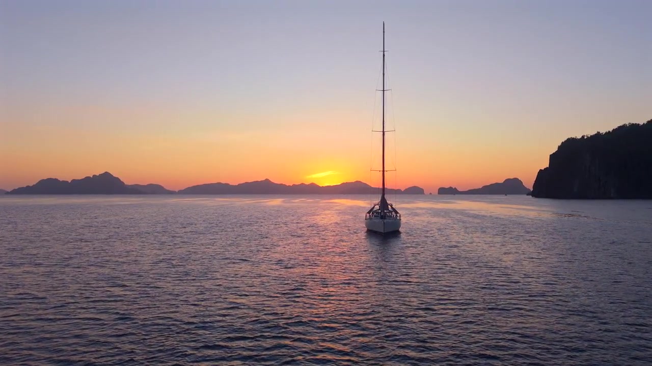 Veleiro centralizado repousa em água texturizada com o sol se pondo atrás das distantes ilhas de El Nido.