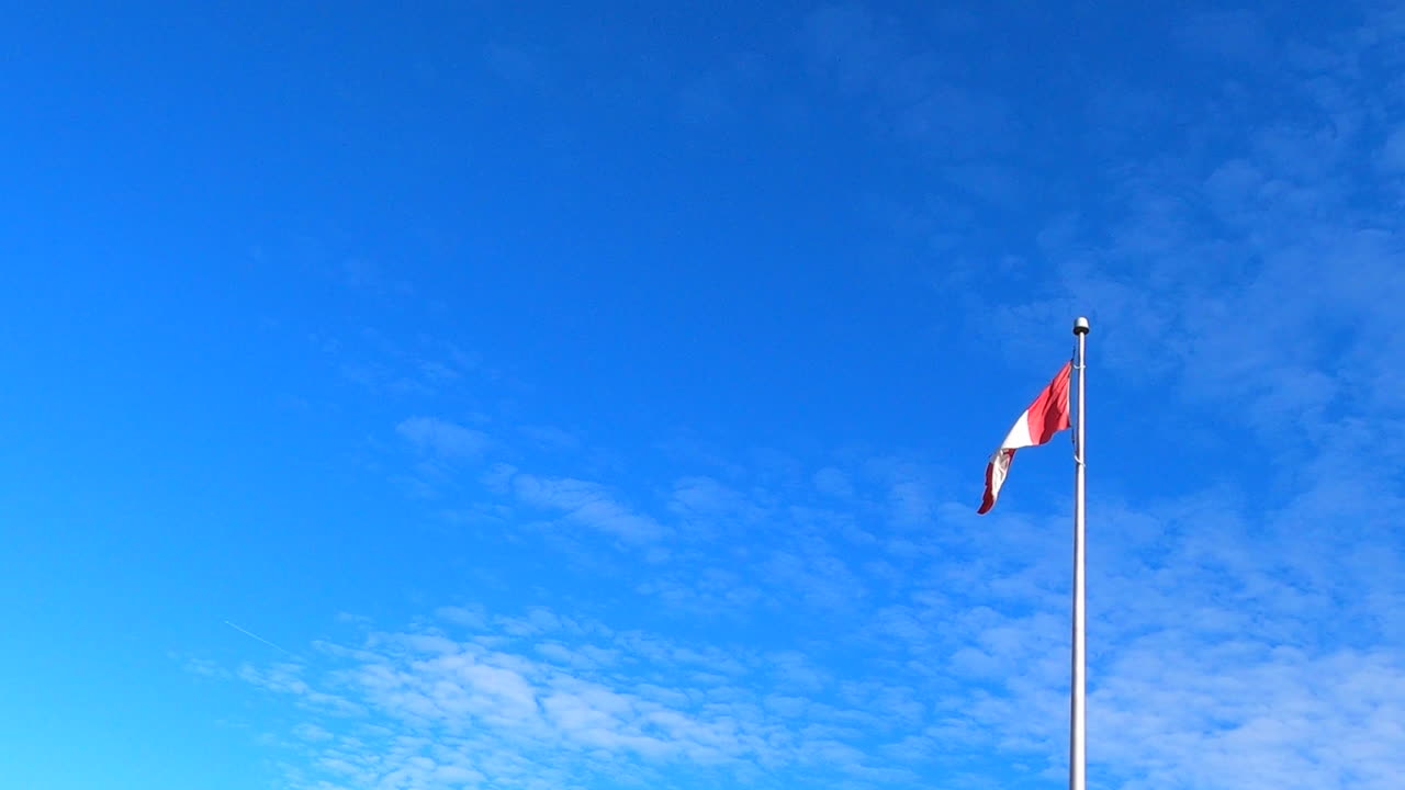 Bandiera nazionale canadese su pennone che sventola, cielo azzurro sullo sfondo, inquadratura dal basso in campo lungo.
