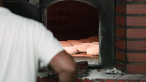 back view of a baker with metal peel putting the kneaded dough inside the hot oven. - close up shot
