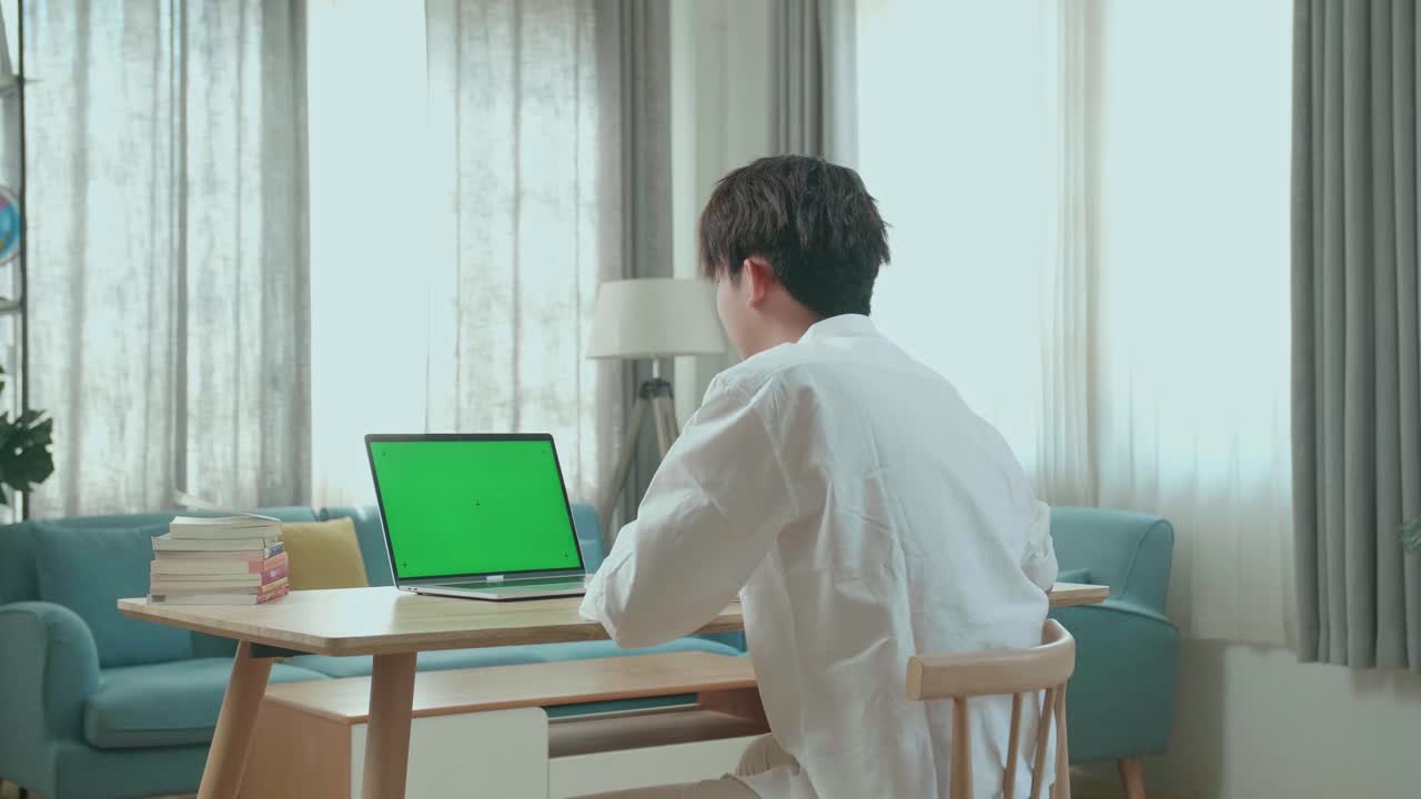 Hind View Of Asian Man Student Raises Hand And Asks A Question While Using Green Screen Computer At Home