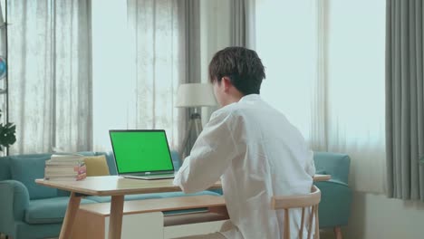 hind view of asian man student raises hand and asks a question while using green screen computer at home
