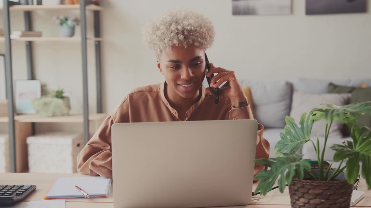 African woman talking on mobile phone while sitting at home office with laptop