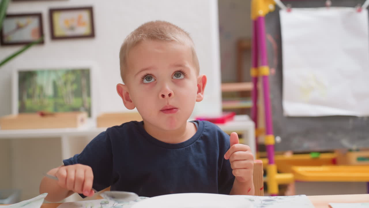 Ragazzino curioso con una maglietta blu scuro che cerca di mangiare prima di prenderlo, guarda avanti con espressione pensierosa mentre una bambina con una maglietta rosa cammina dietro di lui portando un piatto in un'aula ben illuminata con decorazioni educative