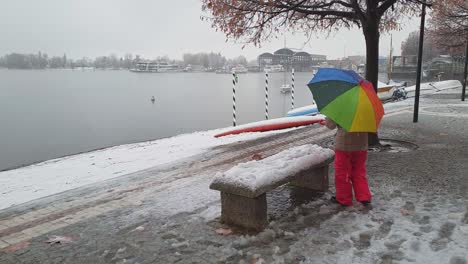 child holding beautiful colorful umbrella plays with snow on bench in arona