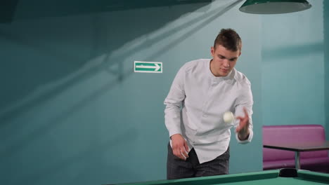 caballero con camisa blanca de pie frente a la mesa de billar, recogiendo la pelota de billar blanca. expresión enfocada, entorno moderno de club deportivo, mesa de billard verde, iluminación aérea
