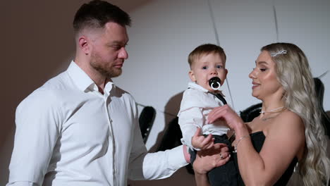 blonde long-haired woman holding a baby boy in white shirt and sucking a pacifier. father stands beside. caucasian family portrait in studio.