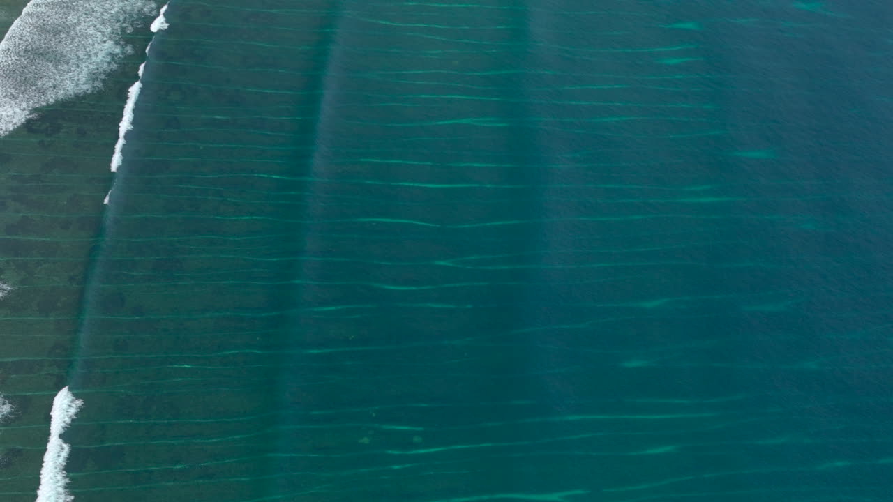 las ondas suaves crean patrones en el océano azul profundo que muestran patrones detallados de las estructuras de arrecife debajo de la superficie del océano con olas blancas rompiendo a lo largo de la costa.