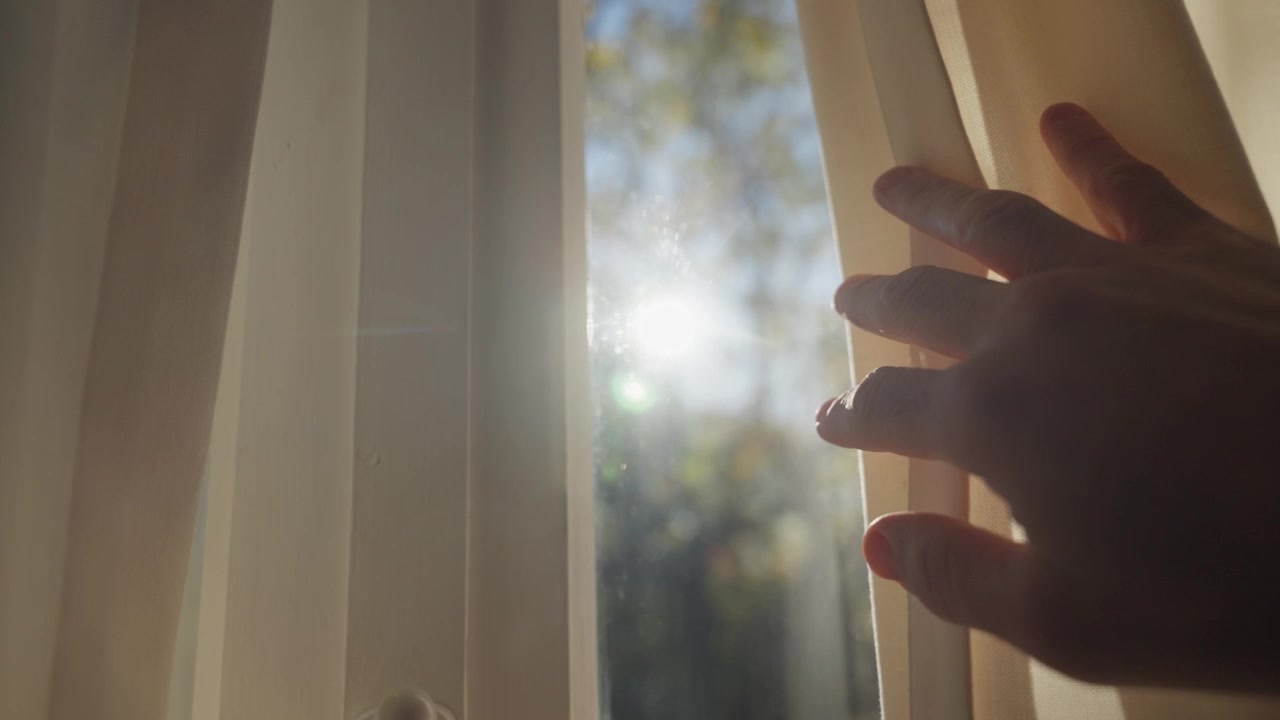 Close up on hand reaching towards window curtain to open and reveal the sun shining inside room in early morning light with green out of focus forest or garden. Symbol of hope and faith. Enlightenment