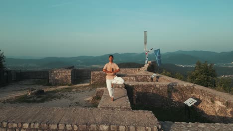 Drone-shot-of-Indian-man-standing-in-hatha-yoga-pose-standing-on-one-leg-on-stone-castle-wall-on-top-of-the-hill-surrounded-by-hills-fields-and-forests-in-the-morning-at-sunrise