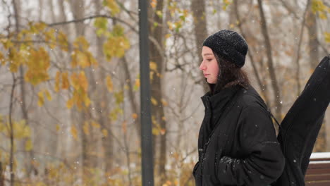 young girl in winter jacket with hands in pockets and guitar on back walking in snowfall, snowy park bench and colorful autumn leaves in background