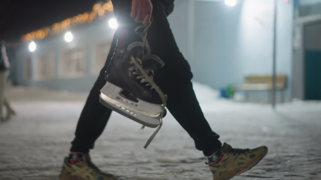 Close up of skater walking outdoors holding black ice skating boot by lace, with background showing another skater in soft blur carefully moving on icy ground at night near lighted winter building