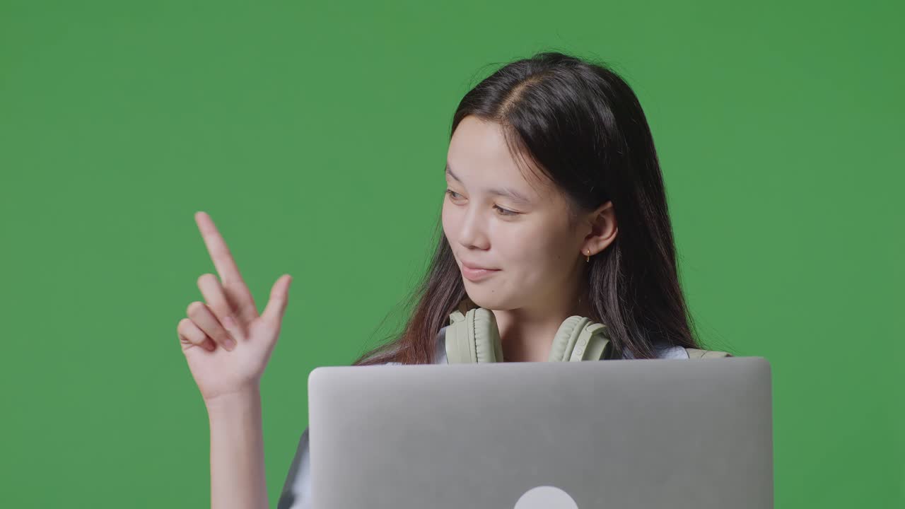 배을 들고 있는 아시아 십대 소녀 학생의 가까운 사진. 녹색 화면 배경 스튜디오에 서 있을 때 으로 가리키고 노트북을 사용합니다.