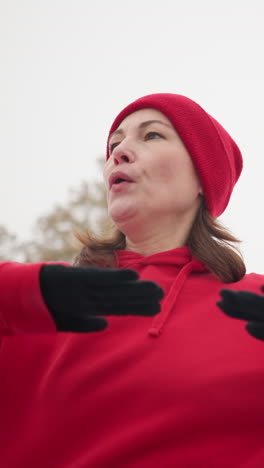 close-up of lady exercising outdoors raising hands for balance turning left and right in winter setting with snowy hill iron railing blurred light pole vibrant red hoodie for warmth visible breath