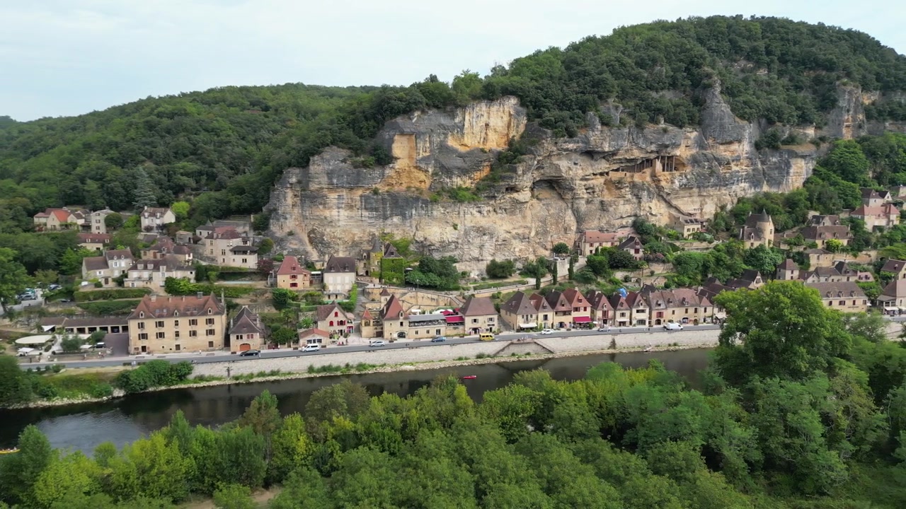 Veduta aerea con drone nella campagna francese di una piccola e antica città medievale sotto una scogliera di pietra, circondata da una foresta verde e da un fiume che la circonda a La Roque Gageac