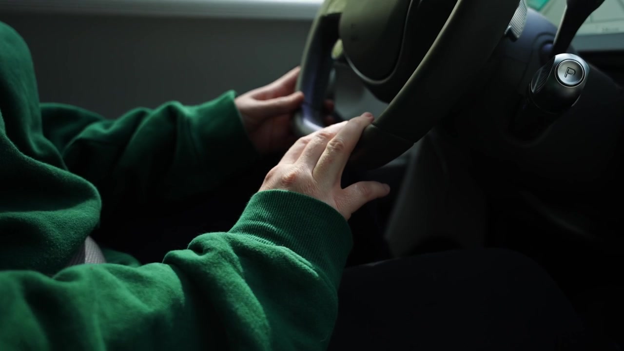 Primer plano de las manos de un conductor en el volante dentro de un coche, con suéter verde, detalle del salpicadero y ambiente de conducción diario tranquilo