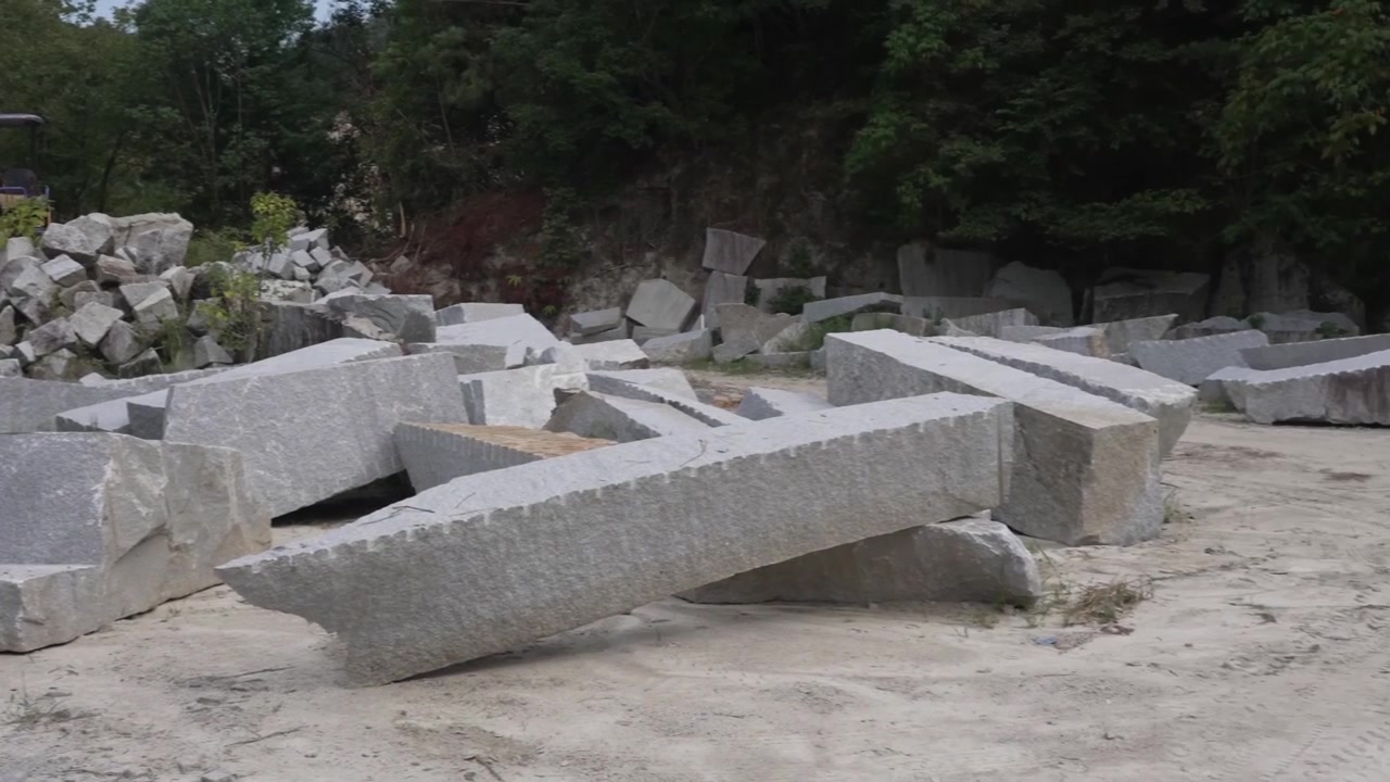 Lange granitplader og afskårne blokke ligger spredt på en japansk stenbrudsplads med en gravemaskine parkeret ved den skovklædte bjergside
