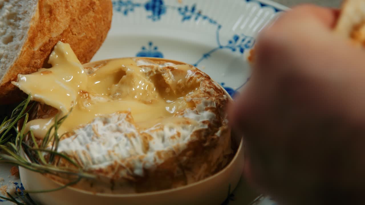 Two Hands Dipping Baguette in Baked French Camembert with Rosemary on Plate
