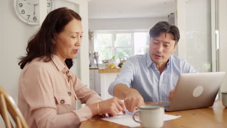 mature asian couple sitting at table at home using laptop to organise household bills and finances - shot in slow motion