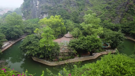 view from ngoa long mountain of a traditional pavilion surrounded by tranquil emerald pond waters, with dramatic limestone cliffs rising in the background, at hang mua resort in ninh binh.
