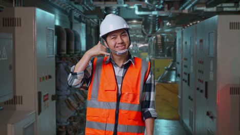asian male engineer with safety helmet smiling to camera and making call me gesture while standing in engine control room, work of electrical generators