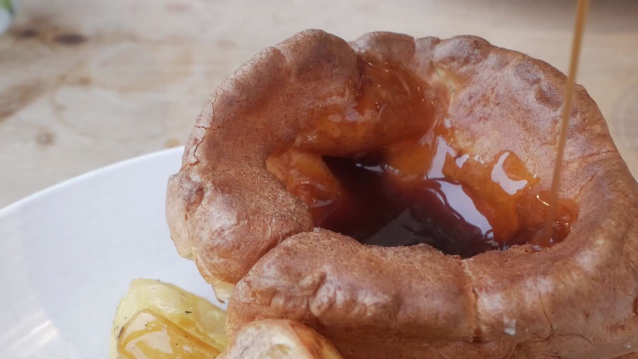 A pour of gravy into a freshly baked Yorkshire pudding, with roasted potatoes on the side