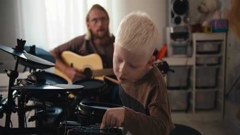 an albino boy with white hair sets up electric drums to start playing his favorite tunes along with his father, a blond man in glasses with a beard. happy before musicians, a blond man with a beard plays an acoustic guitar and his little son plays along with him on the drums