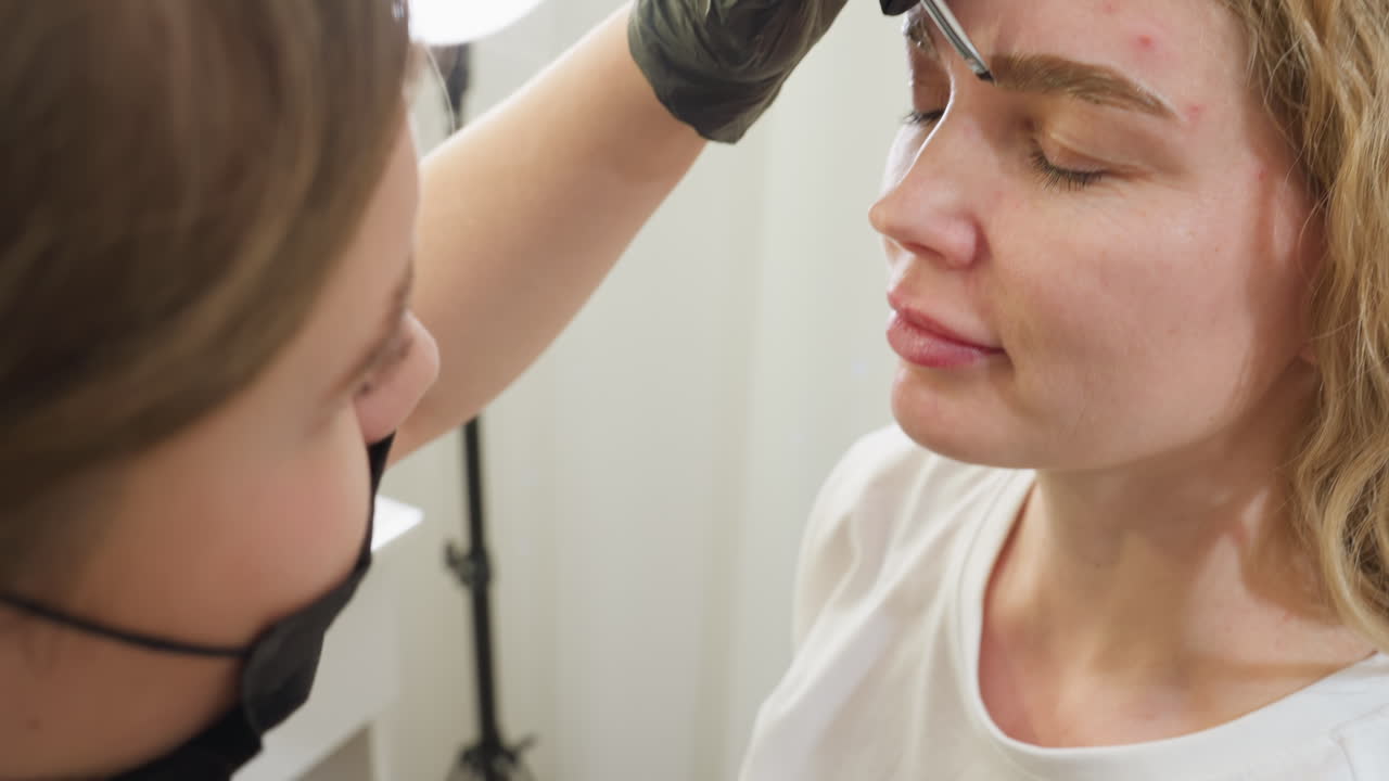 Beautician wearing black gloves holds small glass jar containing cream used for eyebrow treatment while preparing to apply on client. Blurred background shows part of client and ring light stand